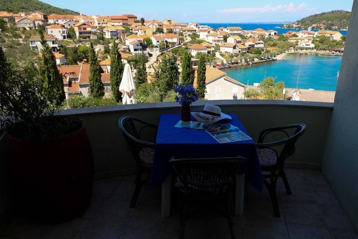 A balcony with a table set for two, overlooking a coastal town with houses and a body of water.