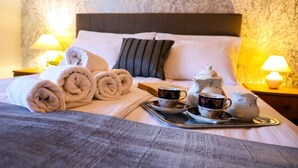 Una cama bien hecha con toallas enrolladas, una bandeja con tazas de té y una tetera, y una mesita de noche con una lámpara.