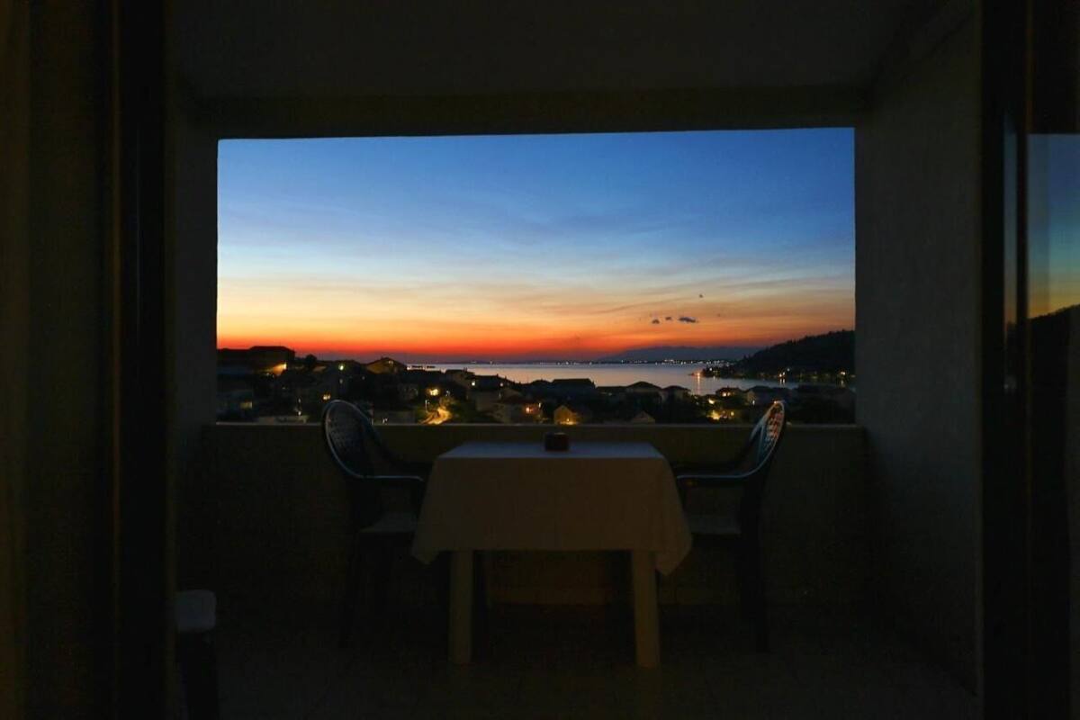 A balcony with a table and chairs, overlooking a sunset view of a cityscape and water.