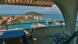 A balcony with a table and chairs, overlooking a marina with boats and a coastal town. - Apartments Novis (Kali)