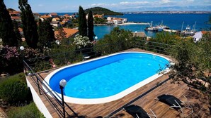 Ein Pool mit Holzterrasse und Liegestühlen, umgeben von Grünflächen mit Blick auf eine Küstenstadt und einen Hafen mit Booten.