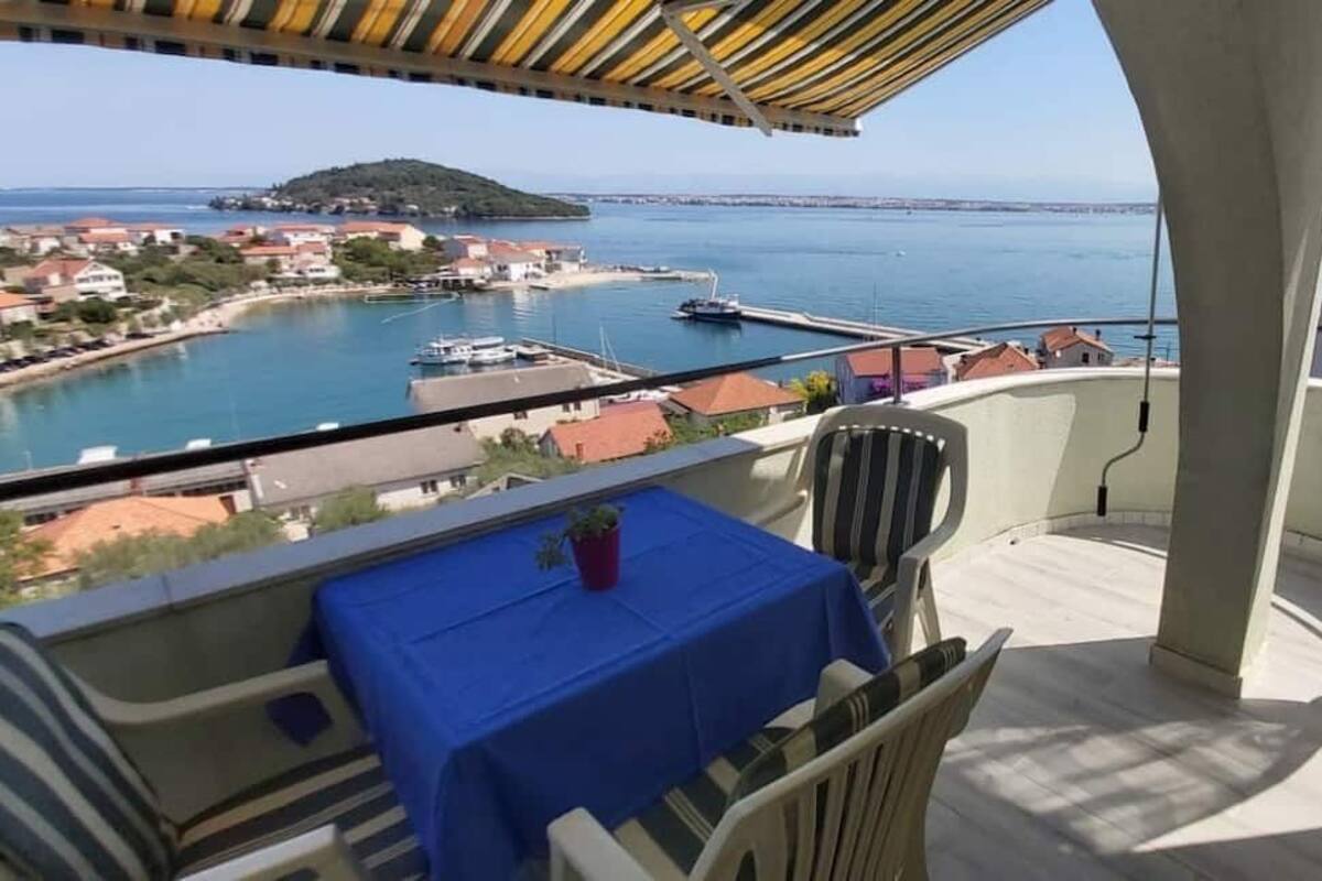A balcony with a table and chairs, overlooking a coastal town and the sea.