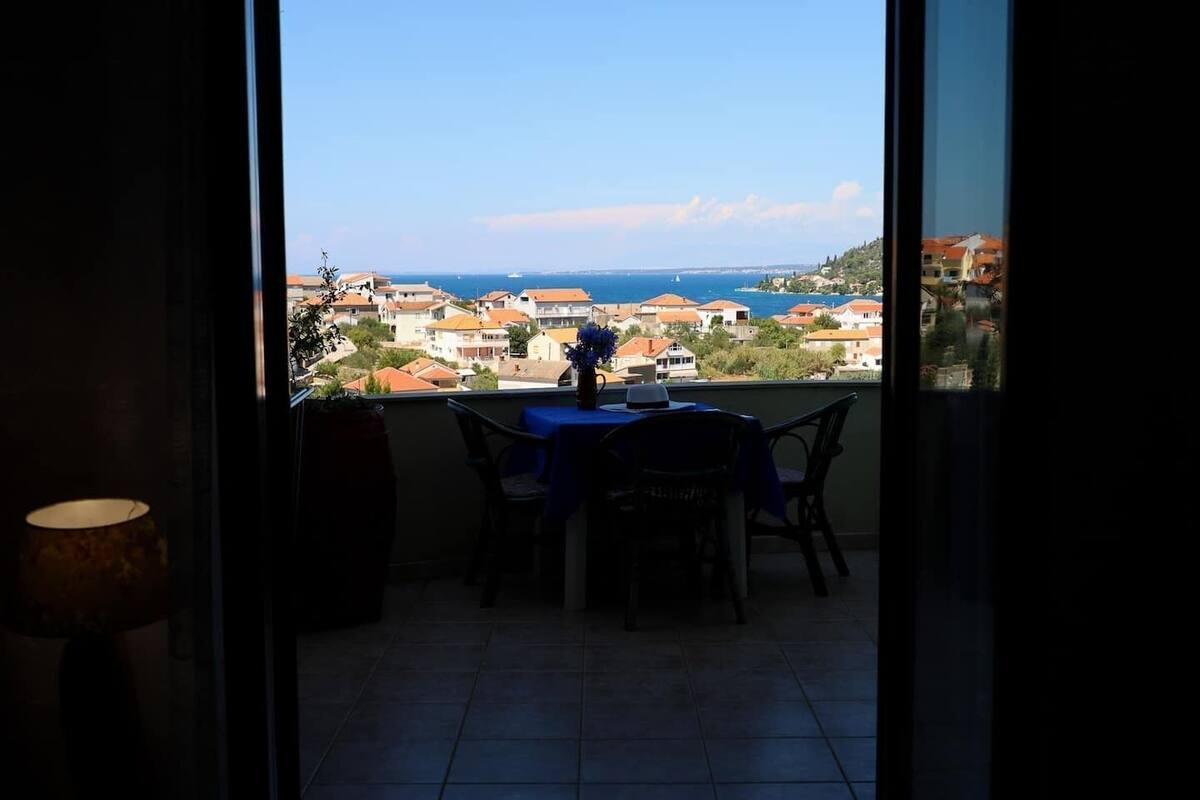 A balcony with a table and chairs, overlooking a coastal town with houses and the sea.