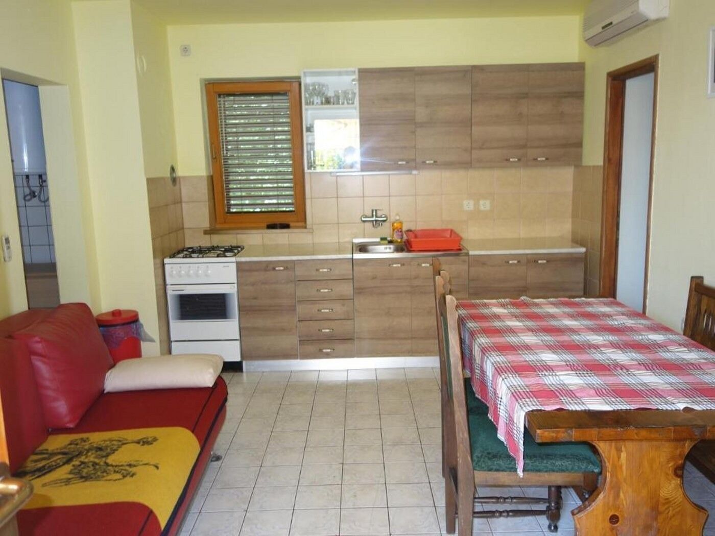 A compact kitchen with a stove, sink, and wooden cabinets. There is a red sofa and a wooden dining table with a checkered tablecloth.