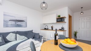 Uma sala de estar moderna com um sofá cinzento, uma mesa de jantar com uma tigela de frutas e uma garrafa de vinho, e uma área de cozinha com armários brancos e um micro-ondas.