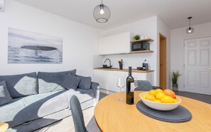 A modern living room with a grey sofa, a dining table with a bowl of fruit and a wine bottle, and a kitchen area with white cabinets and a microwave. - Apartments Marijo (Baska Voda)