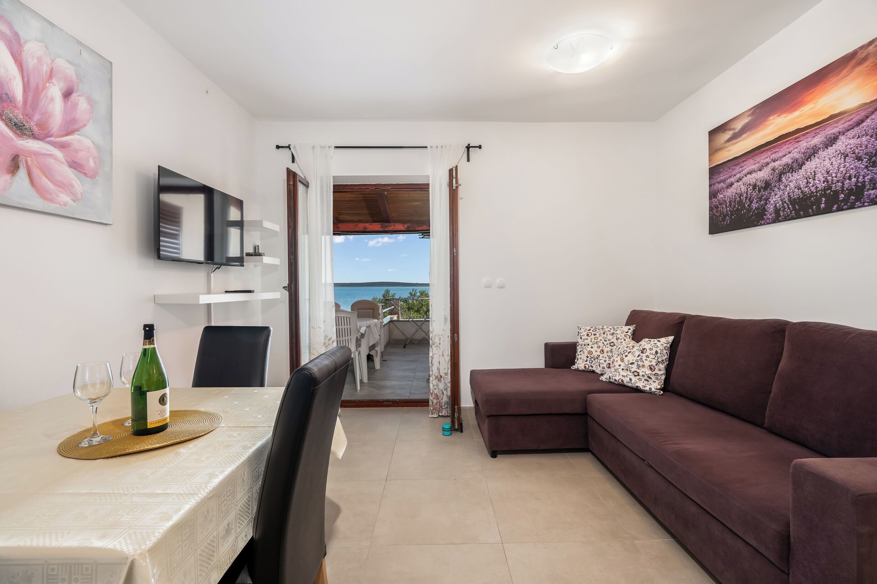 A living room with a brown sofa, a dining table with a bottle of wine, and a TV on the wall.