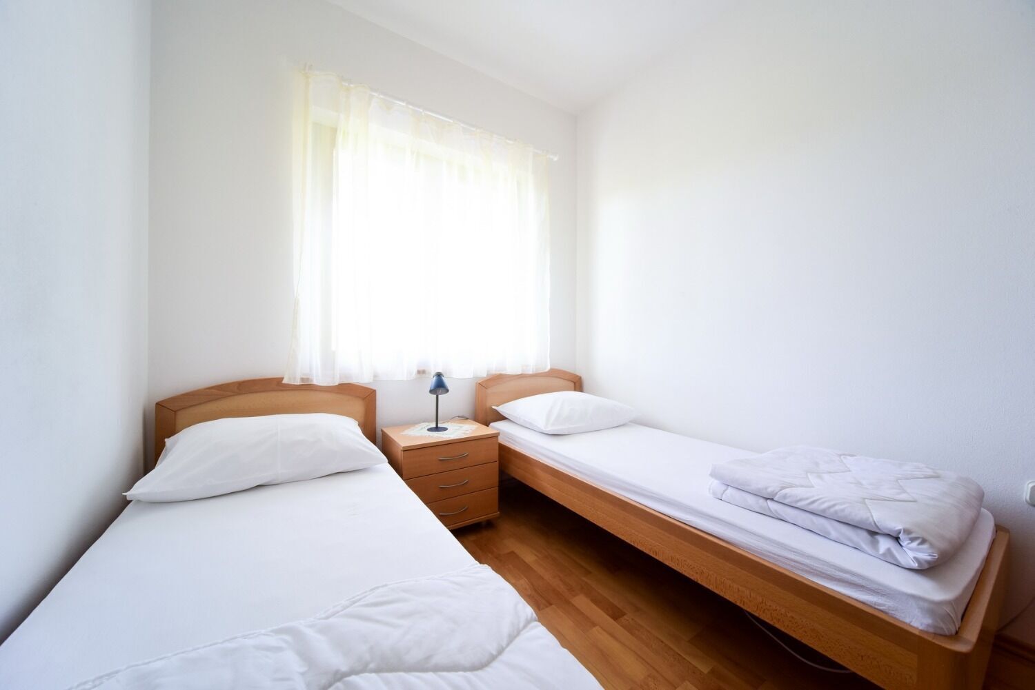 Una stanza piccola e pulita con due letti singoli, un comodino, una lampada e una finestra con tende leggere.