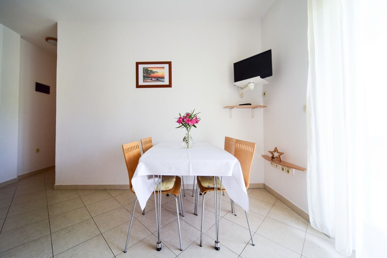 A dining area with a white table, four chairs, a vase with flowers, a framed picture on the wall, a mounted TV, and a wooden shelf with a star decoration.