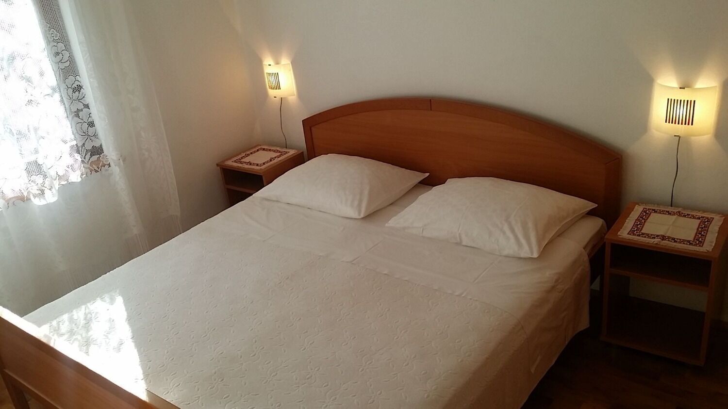 A neatly made bed with two pillows, wooden headboard, bedside tables with lamps, and lace curtains.