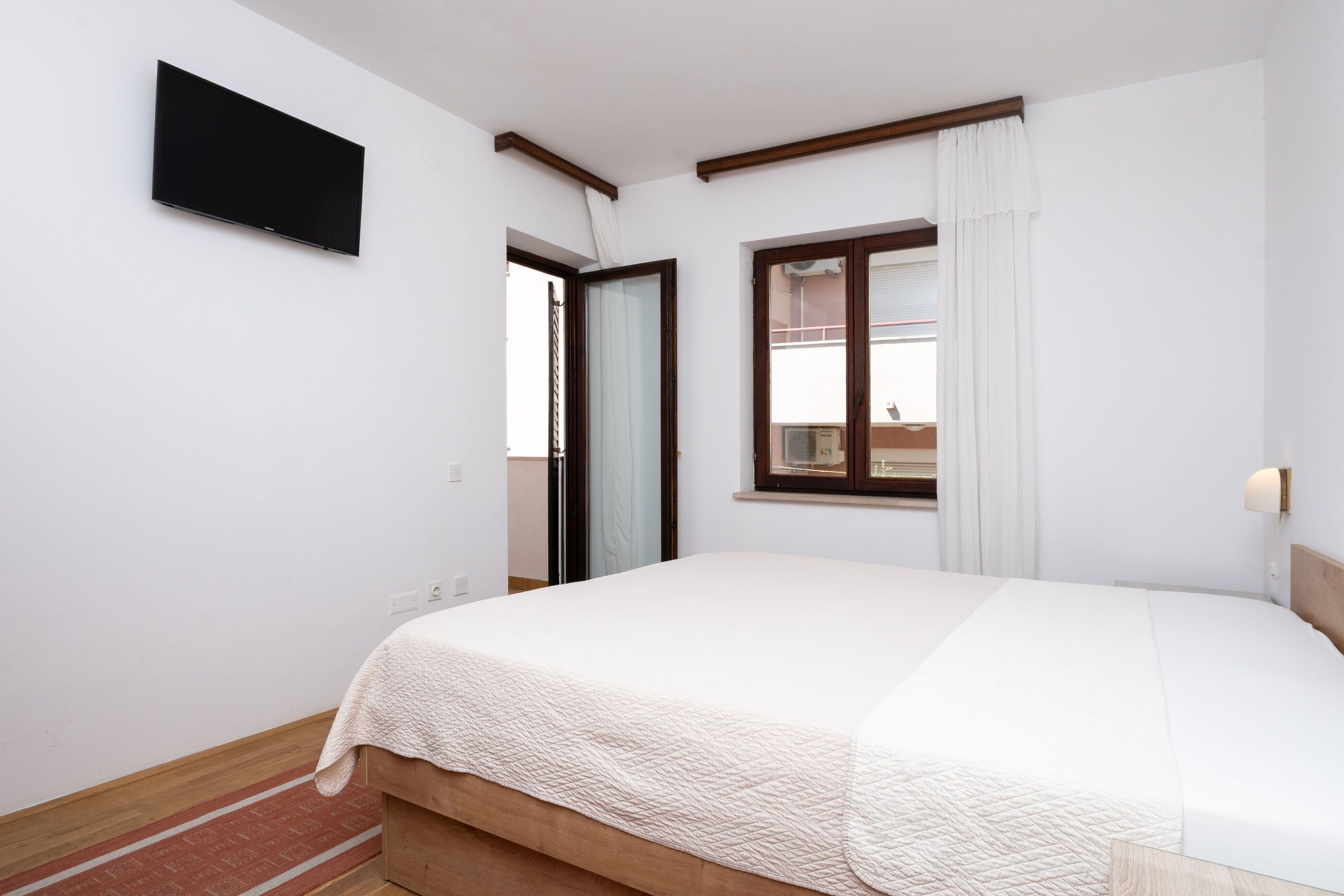 Un dormitorio con cama, televisor montado en la pared, ventana con cortinas y puerta que da a un balcón exterior.