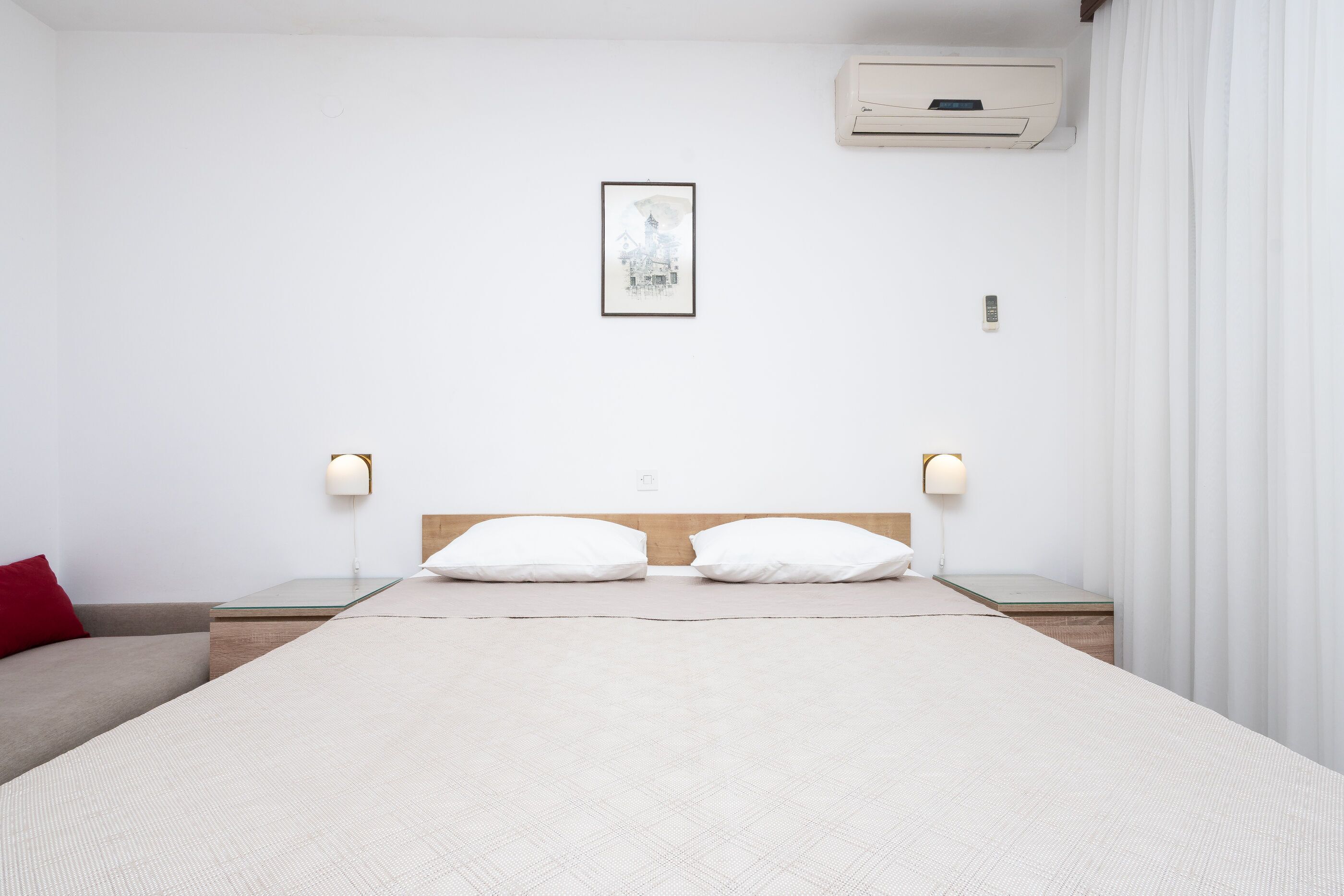 Une chambre d’hôtel comprenant un lit, deux tables de chevet, un climatiseur fixé au mur, un tableau encadré et une fenêtre avec des rideaux.
