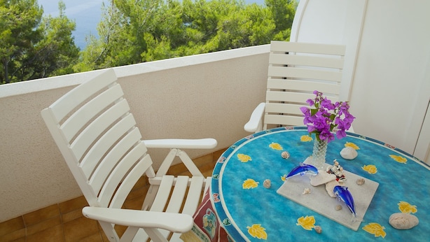 Un balcone con sedie bianche, un tavolino con tovaglia blu e un vaso con fiori viola.
