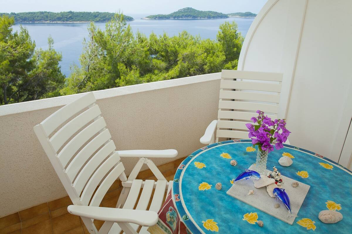 Un balcone con sedie bianche, un tavolino con tovaglia blu e un vaso con fiori viola.