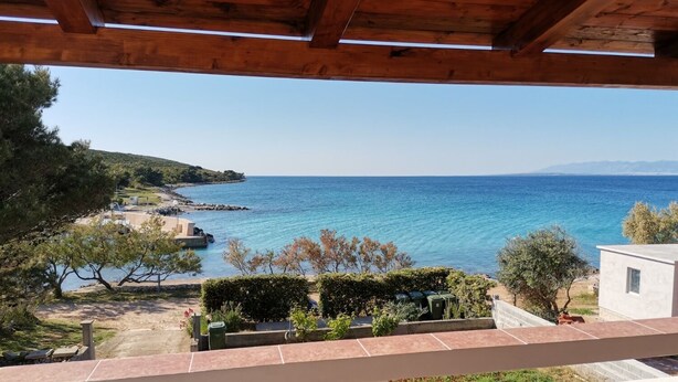 Vista costeira a partir de uma varanda com teto de madeira, com vista para um mar azul límpido e uma linha costeira rochosa.