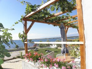 En træpergola med slyngplanter, et bord med stole og lyserøde blomster i en have ved vandet.
