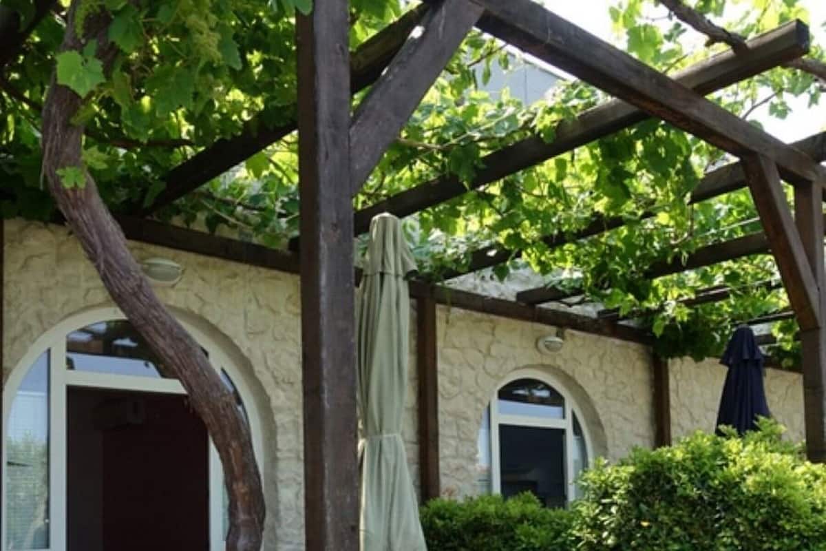 A covered outdoor seating area with a table and chairs, a pergola structure, and a stone wall.