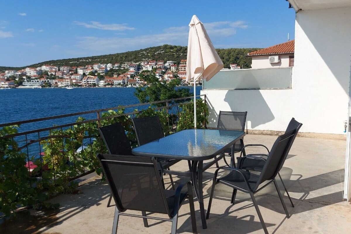 A balcony with a glass table and chairs, overlooking a coastal town and the sea.