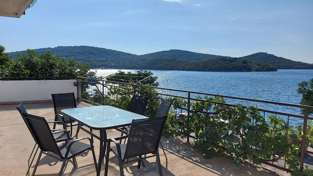An outdoor terrace with a table and chairs, overlooking a lake with mountains in the background. - Apartment Bila (Tisno)