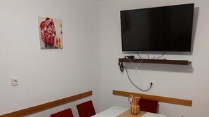 A room with a white table, red chairs, a TV mounted on the wall, and a patterned carpet. - Apartment Bila (Tisno)