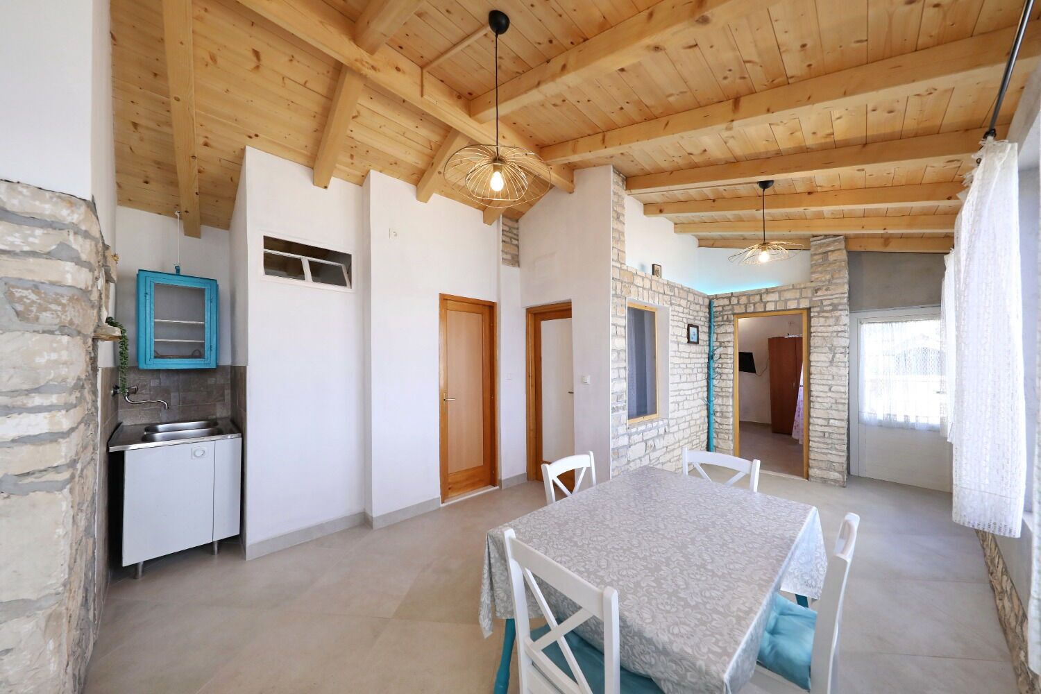 A spacious room with a wooden ceiling, stone walls, a dining table with chairs, a kitchen area with a sink, and a stone fireplace.