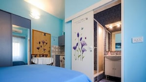 Un dormitorio con una cama, una mesita y una silla. Se ve un baño por una puerta abierta.