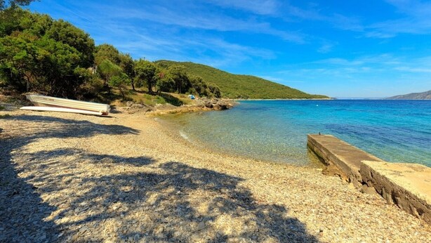 A pebble beach with a boat, clear blue water, and a forested hill in the background. - Apartments Mari (Korčula)