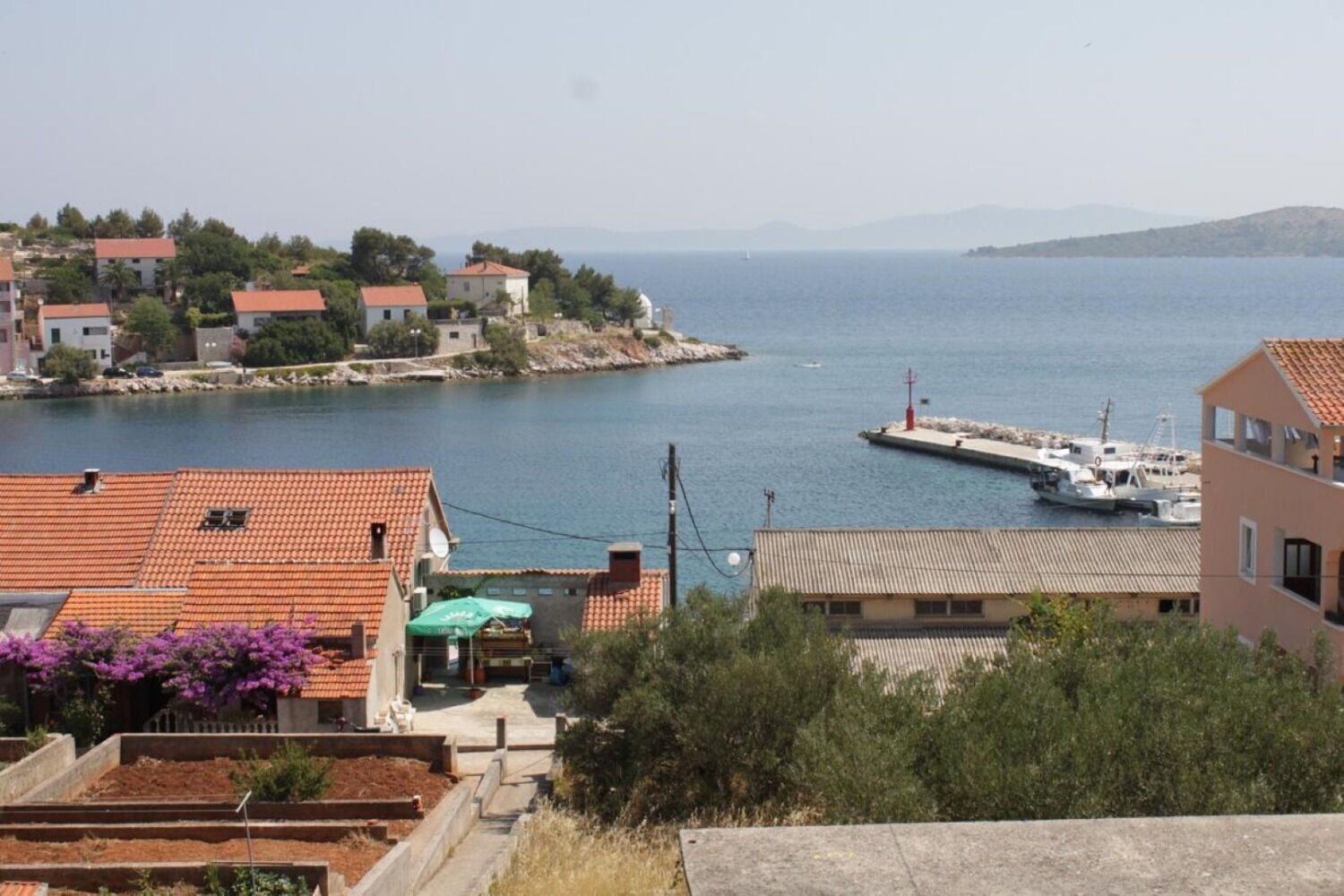 A coastal residential area with terracotta-roofed houses, a dock with boats, and a clear view of the sea.