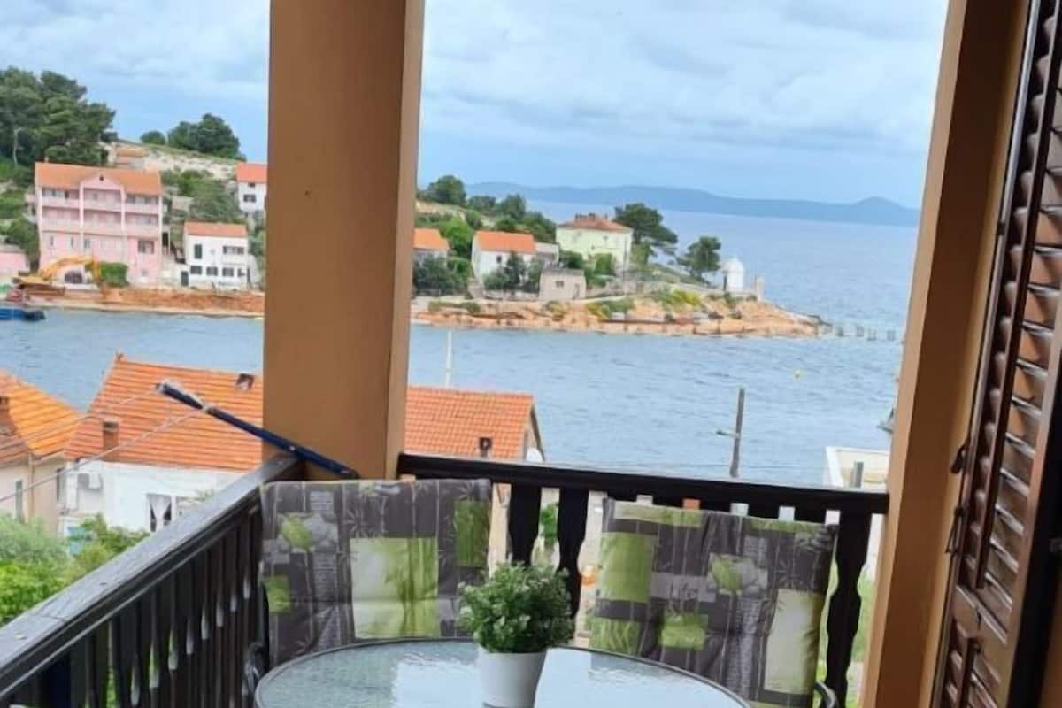 A balcony with a round glass table and two chairs, overlooking a coastal area with buildings and water.