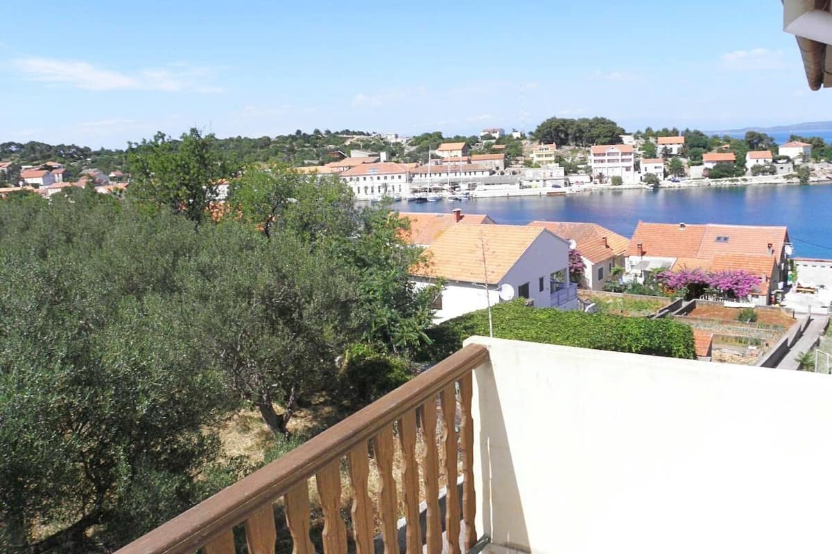 A coastal town with buildings near a body of water, a balcony with a railing, and trees in the foreground.