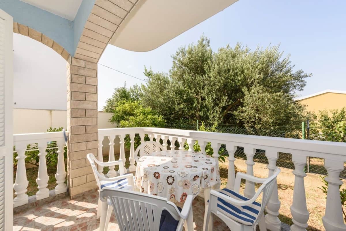 A covered outdoor seating area with a table and chairs, surrounded by a white balustrade and a view of greenery.