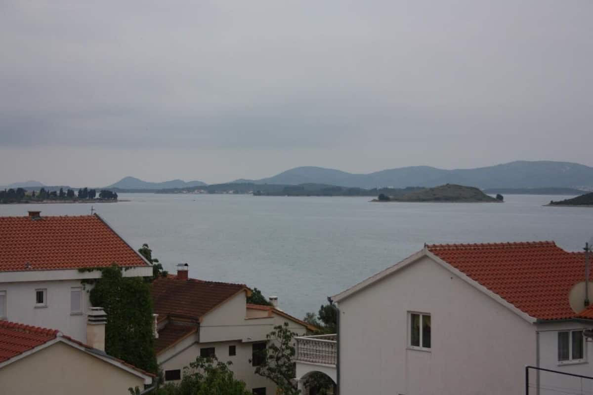 A coastal residential area with houses featuring red-tiled roofs and white walls, overlooking a calm sea with distant hills.