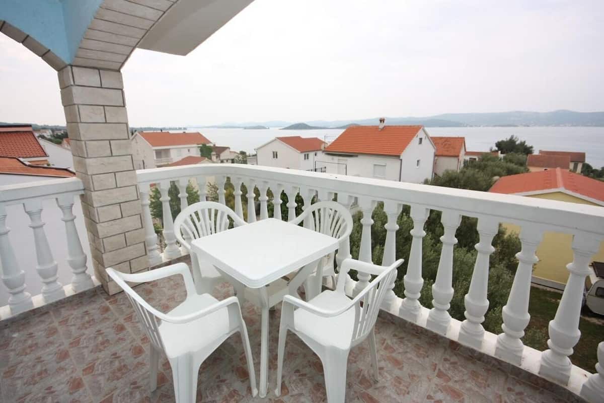 A balcony with a white table and chairs, a white balustrade, and a view of houses and water in the background.