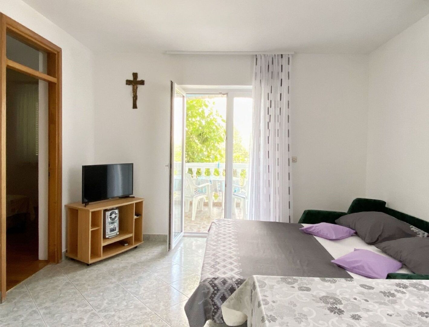 A bedroom with a bed, a TV on a wooden stand, a cross on the wall, and a balcony with a view of greenery.