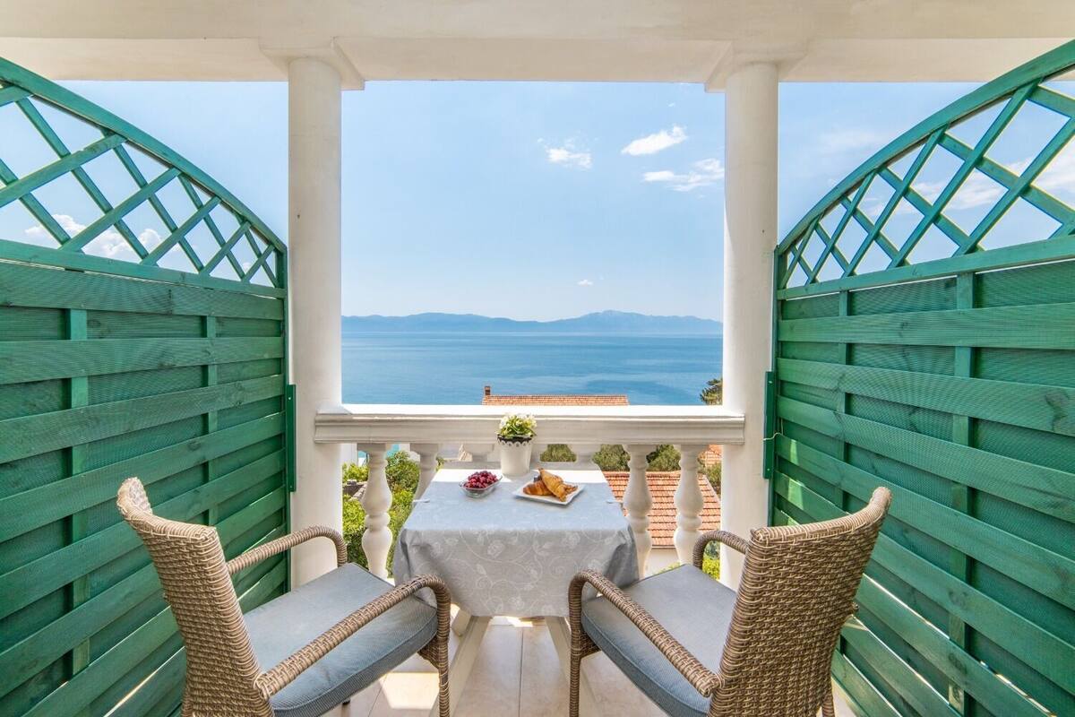 Un balcón con sillas de mimbre y una mesa, con vistas panorámicas al mar y las montañas.