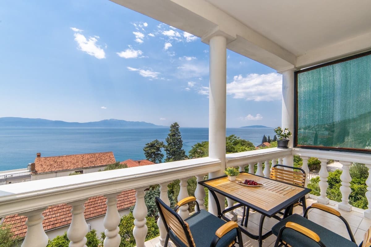Un balcón con mesa y sillas, con vistas al mar y montañas a lo lejos.