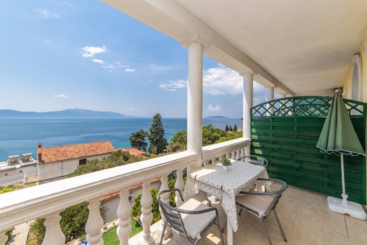 Un balcón con mesa y sillas, una sombrilla verde y vistas al mar y a las montañas.