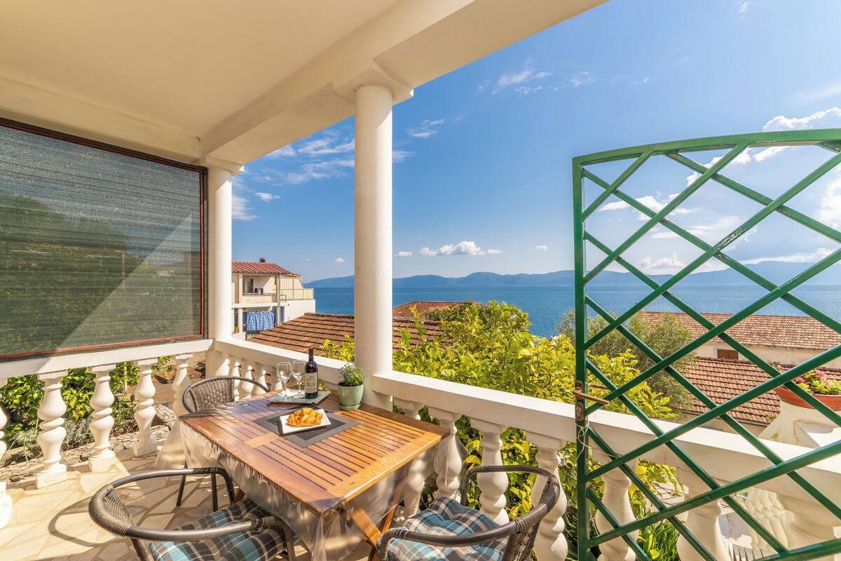 Un balcón con mesa y sillas de madera, vista al mar y barandilla de metal verde.