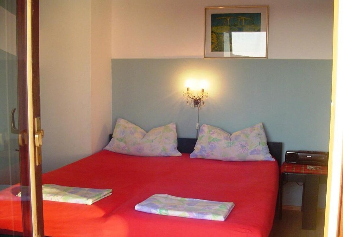 A double bed with red bedding, two pillows, a bedside table with a lamp, and a framed picture on the wall.