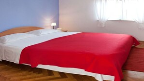 A bed with a red bedspread, a wooden headboard, a nightstand with a lamp, and a window with white curtains. - Apartments Filip (Okrug)