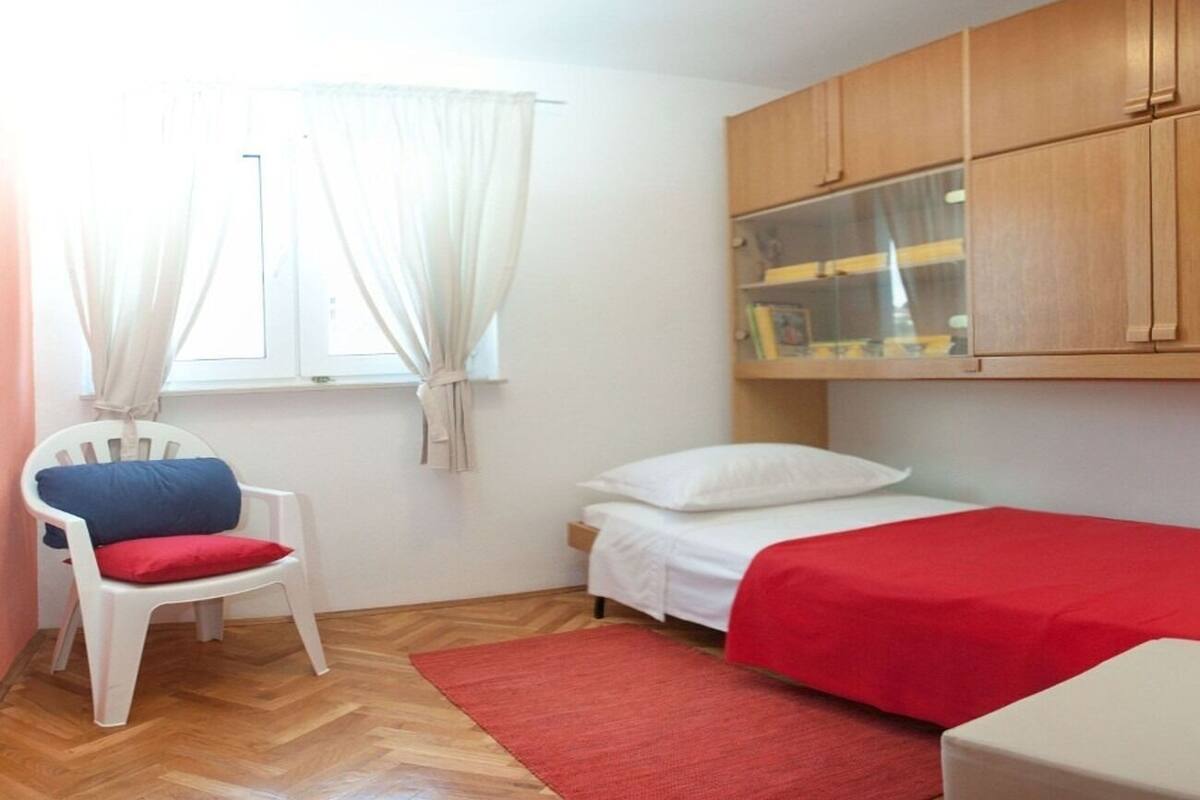 A small, well-lit room with a single bed, a chair with a red cushion, a red rug, and wooden cabinets above the bed.