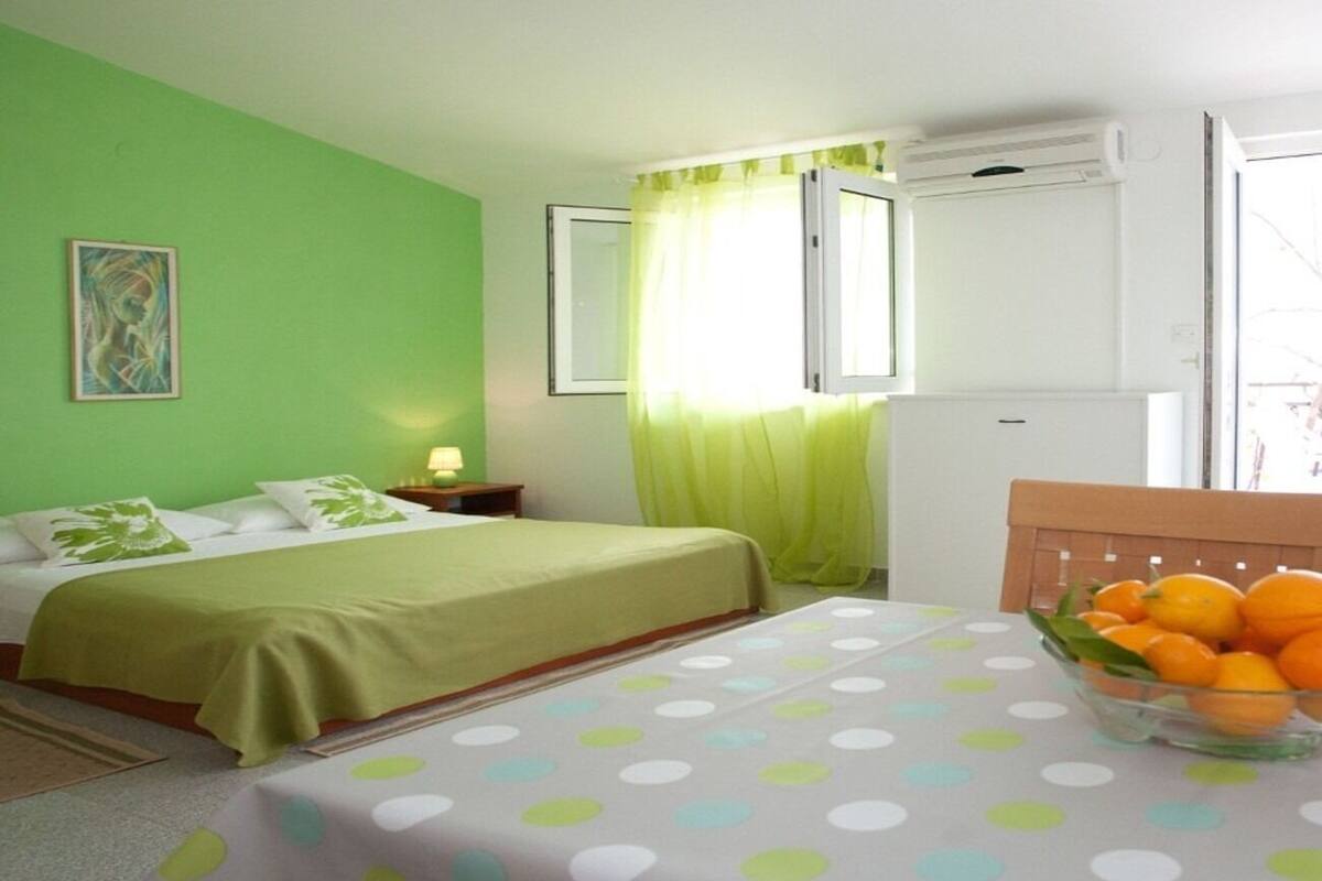 A room with a bed, a table with a fruit bowl, a window with curtains, and an air conditioning unit.