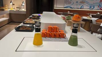 A buffet setup with various food items and beverages, including orange and green drinks, arranged on a white table.
