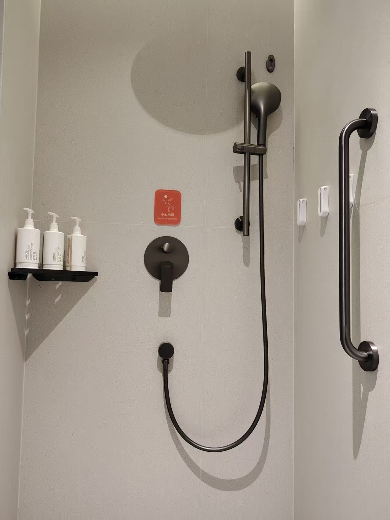 A shower area with a handheld showerhead, a wall-mounted faucet, and a handheld shower hose. There are three bottles of shampoo and conditioner on a shelf.