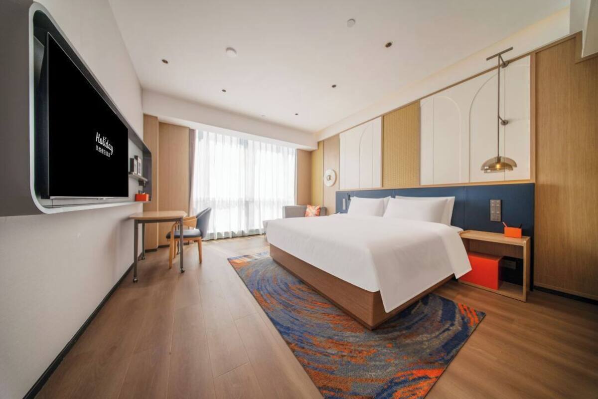 Ein modernes Hotelzimmer mit einem großen Bett, einem Schreibtisch, einem Stuhl, einem Fernseher und einem Fenster mit Vorhängen.