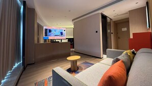 Ett modernt hotellrum med en platt-TV som visar en planritning över rummet, en soffa med kuddar, ett litet runt bord och en hall som leder till en dörr.