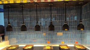 Ett område med buffébord och flera matstationer, med belysning i taket och kakel på väggen bakom.