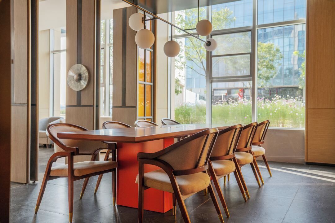 A modern dining area with a large rectangular table surrounded by six wooden chairs, large windows with a view of greenery, and a contemporary light fixture.