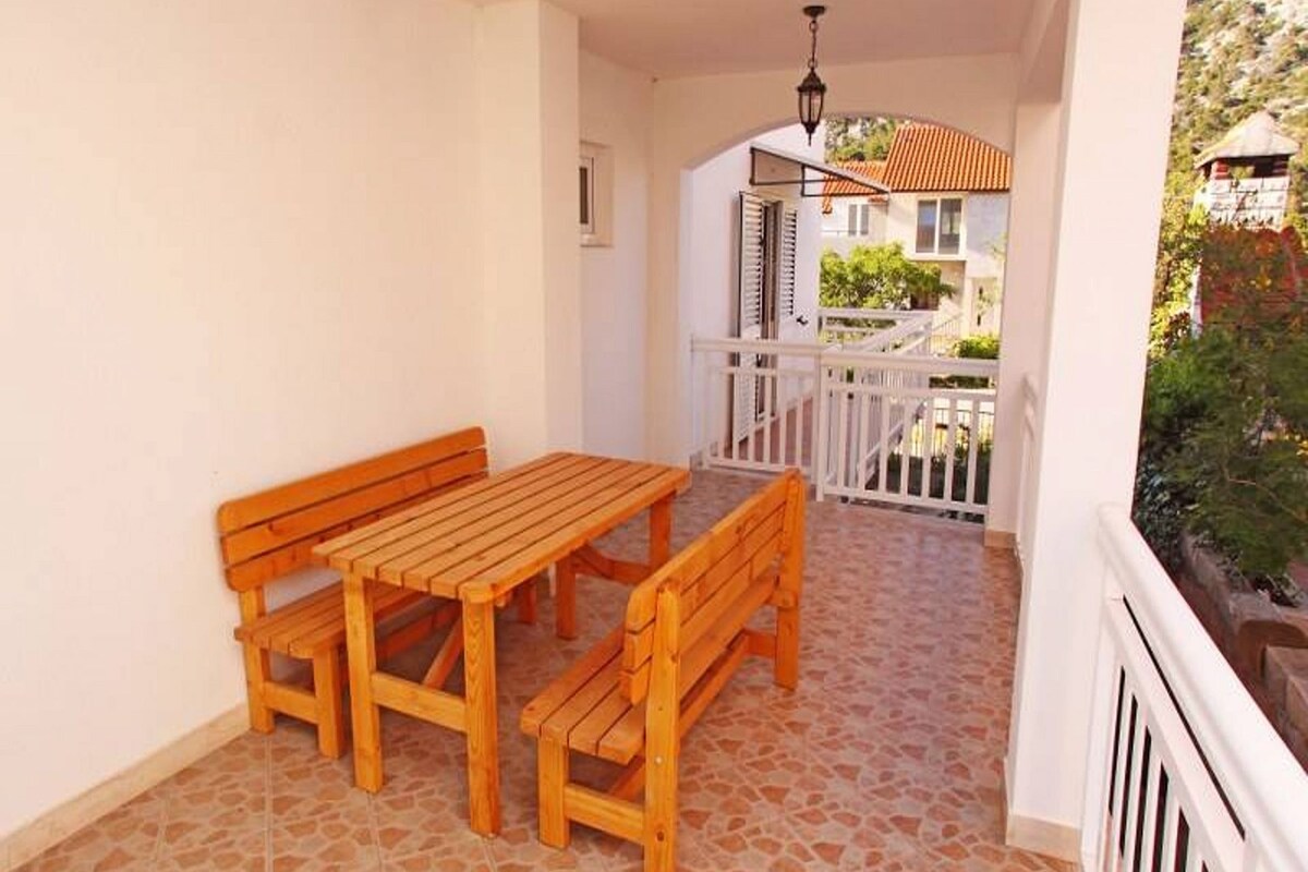 A balcony with a wooden table and benches, white railings, and a view of a house with a red roof.