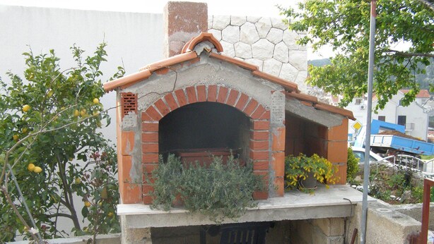 A brick-built outdoor oven with a tiled roof, surrounded by potted plants and a chair. - Apartments Marija (Hvar)